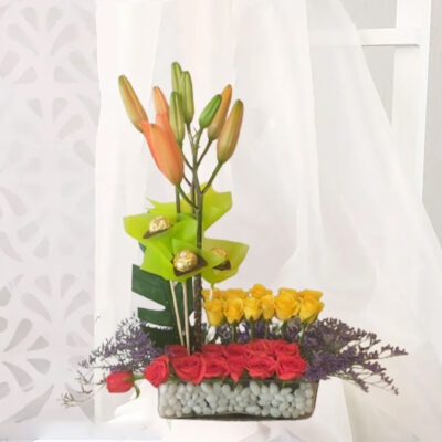 A vibrant floral arrangement featuring 12 Red Roses, 12 Yellow Roses, 3 Ferrero Rocher chocolates, 2 Orange Lilies, and purple flower fillers, accompanied by green leaf fillers, presented in a rectangular 8-inch glass vase with white pebbles.