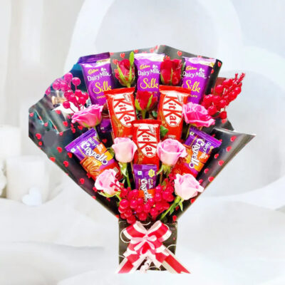 A delightful chocolate assortment including 3 Dairy Milk Silk chocolates (65g each), 3 Dairy Milk Crispello chocolates (50g each), and 3 Nestle Kit Kat chocolates (50g each), accompanied by 4 Baby Pink Roses, 2 Pink Roses, and 2 Red Roses, artificial fillers, red hearts black paper, and a red ribbon bow, presented in a black cardboard box.